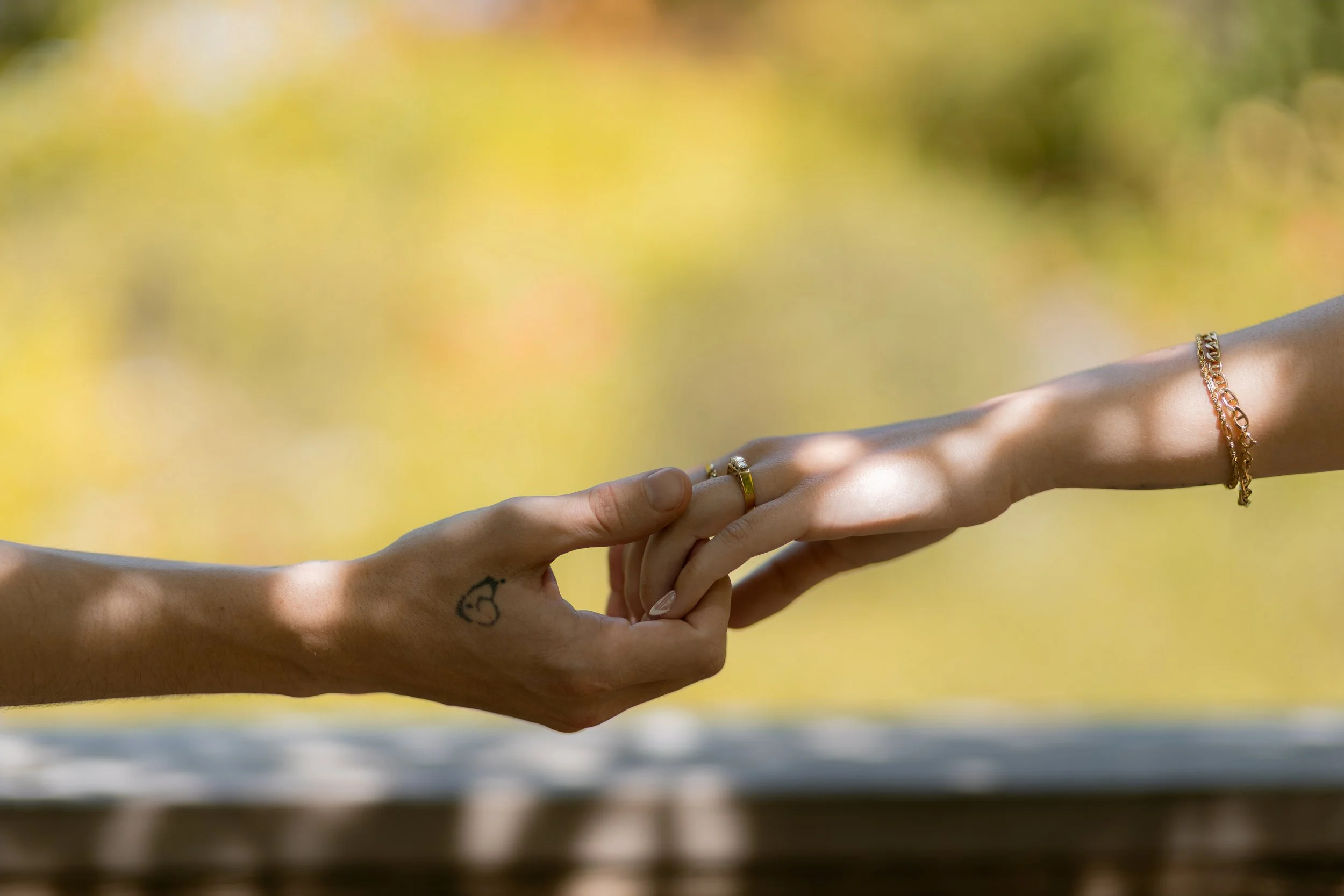 Two hands, one with a tattoo, holding each other's fingers, with blurred yellow and green background.
