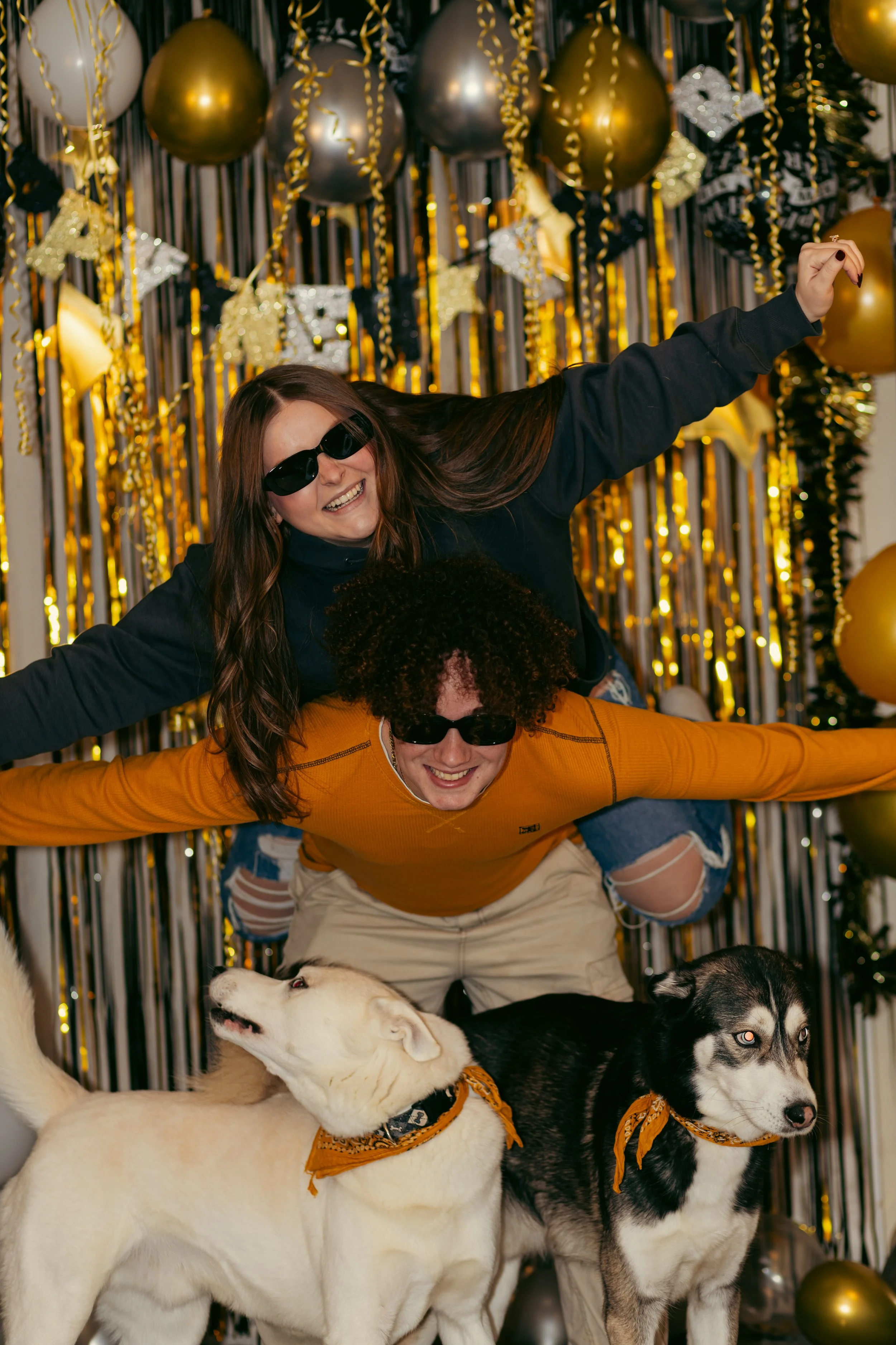 Two people (a woman and a man) with sunglasses, smiling and posing in front of gold and black balloons and streamers, with two dogs wearing bandanas, celebrating at a festive party.