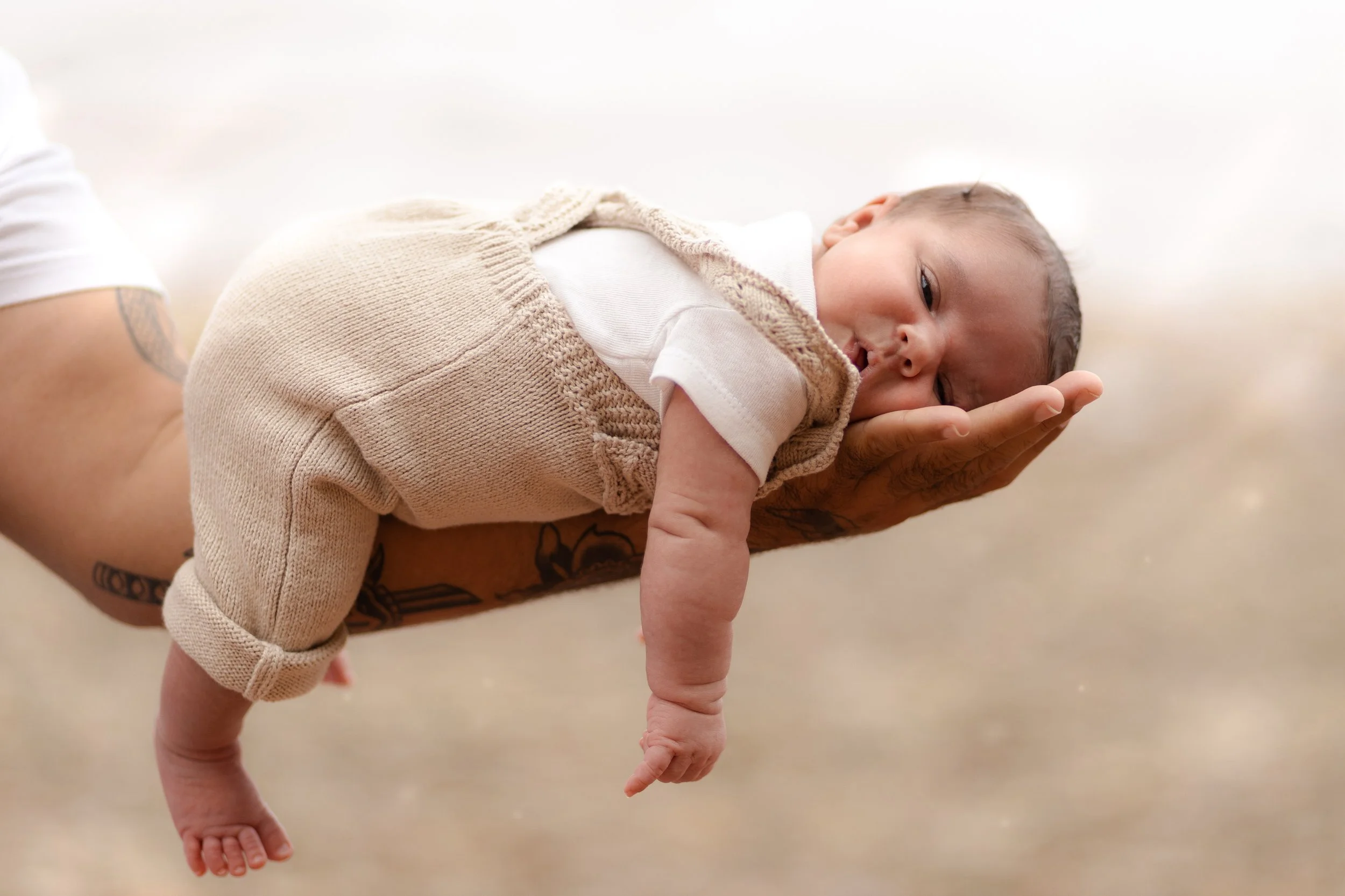 A baby lying on a person's hand, surrounded by a beige knit blanket, with a neutral background.