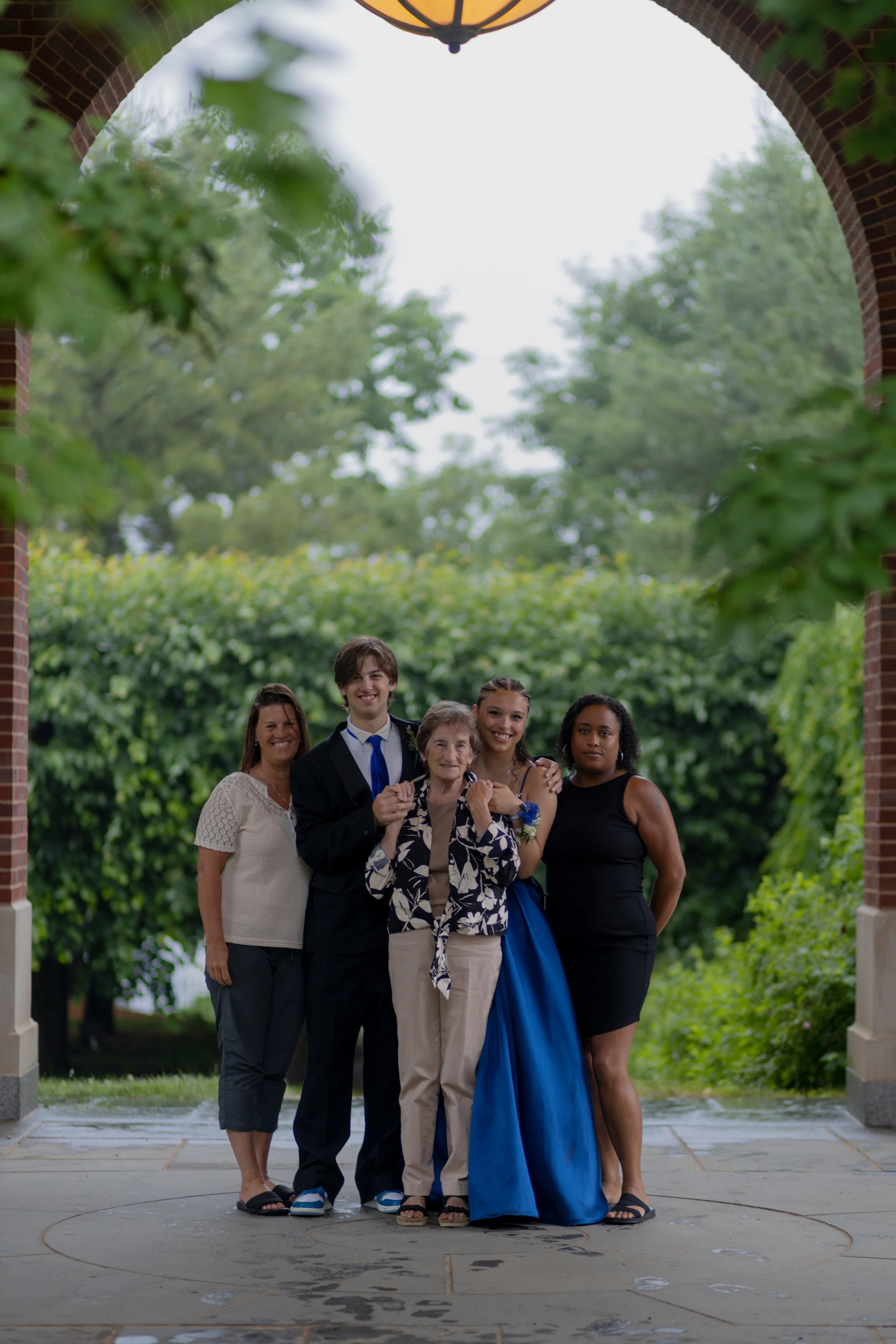 A group of five people standing together under a brick archway during daytime. They are smiling, dressed in formal and semi-formal attire, with a lush green background of trees and bushes.