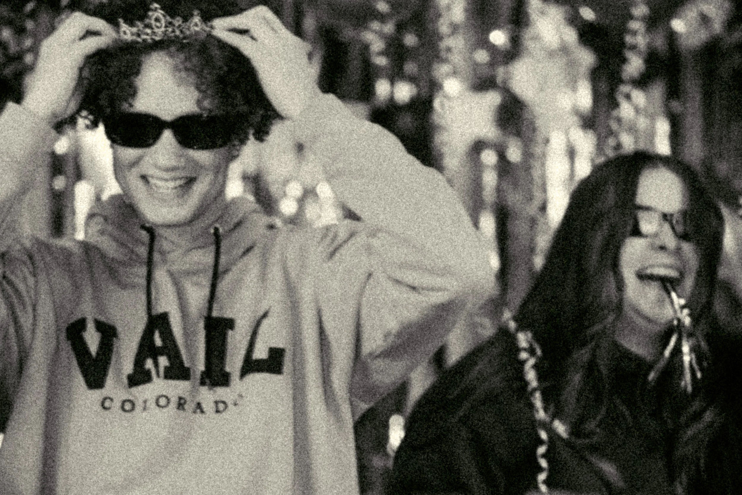 Two women smiling and wearing sunglasses outdoors in a wooded area. The woman on the left is holding a crown or headpiece above her head. The woman on the right has long dark hair and is laughing with her tongue sticking out.