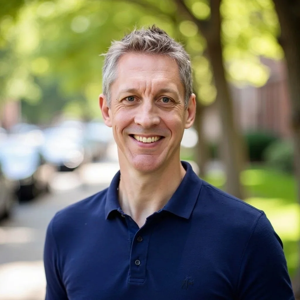 Smiling man with short gray hair, wearing a navy polo shirt, standing outdoors with blurred green trees and parked cars in the background.