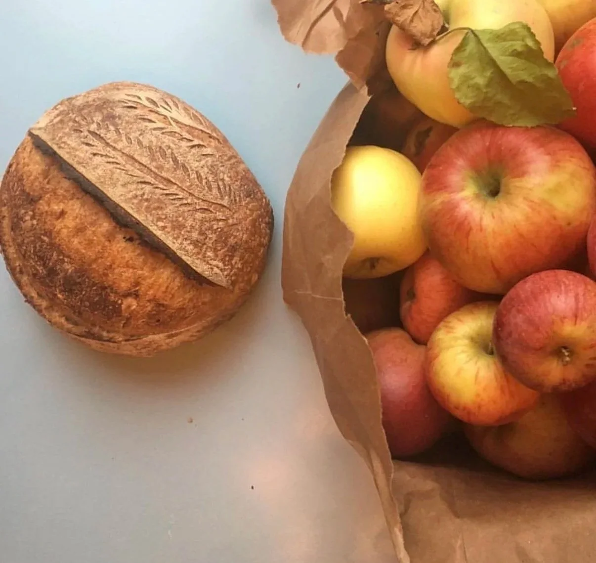 A loaf of rustic bread beside a paper bag filled with red and yellow apples, some with leaves attached.