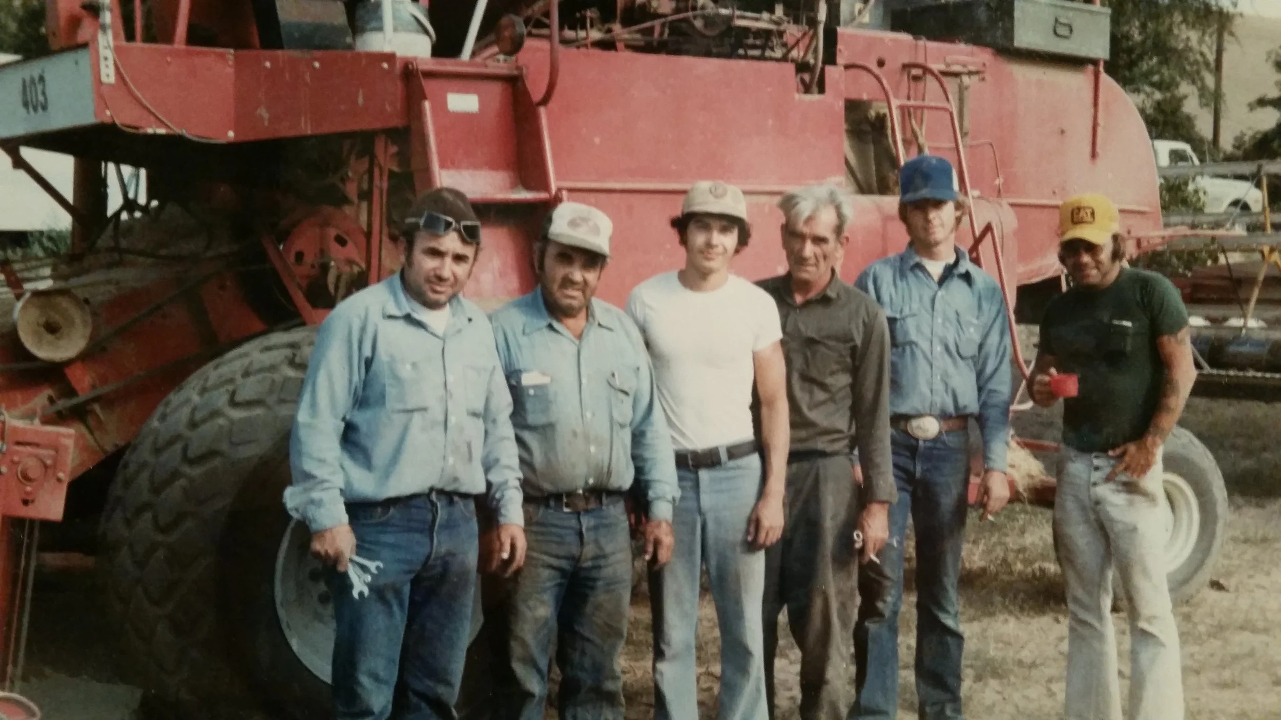 A group of six men standing in front of a large, red piece of farming equipment outdoors, with trees and parked vehicles in the background.