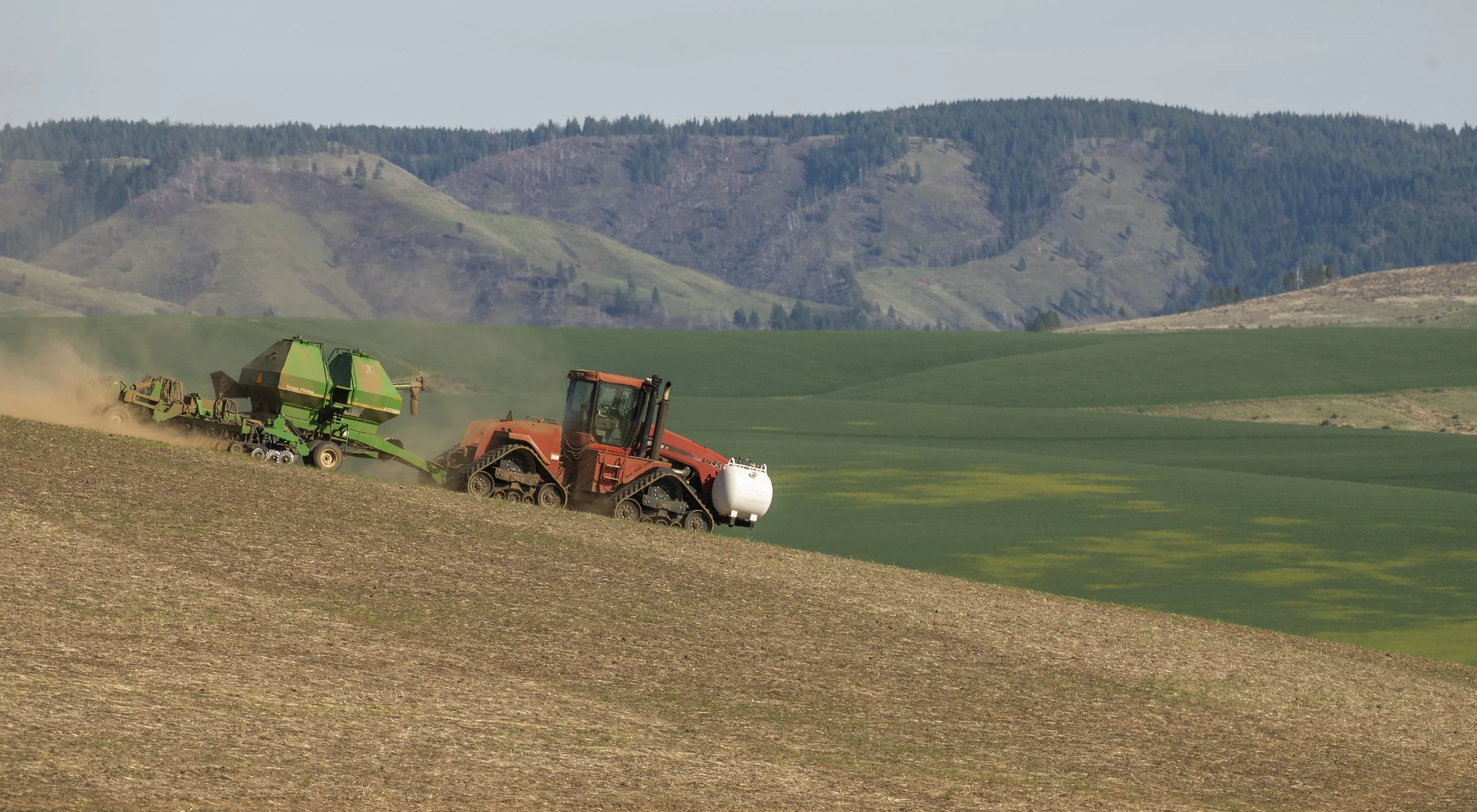 Farm machinery, including a tractor and seed planter, working on a field with rolling hills and mountains in the background.