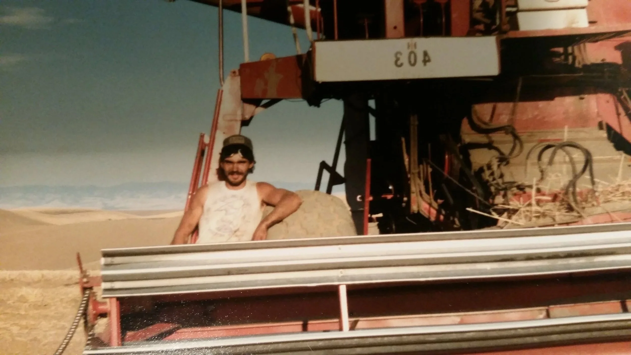 A man with dark hair and a beard, wearing a white sleeveless shirt, standing in front of a vintage steam tractor in a desert setting during sunset.