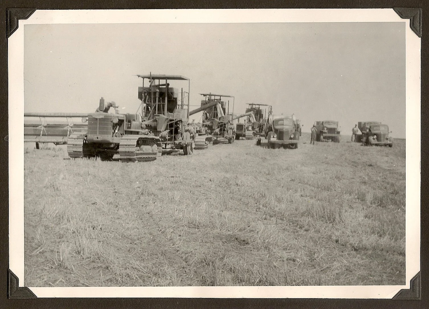 Black and white photo of several vintage tractors and trucks in an open field, with a few people standing around.