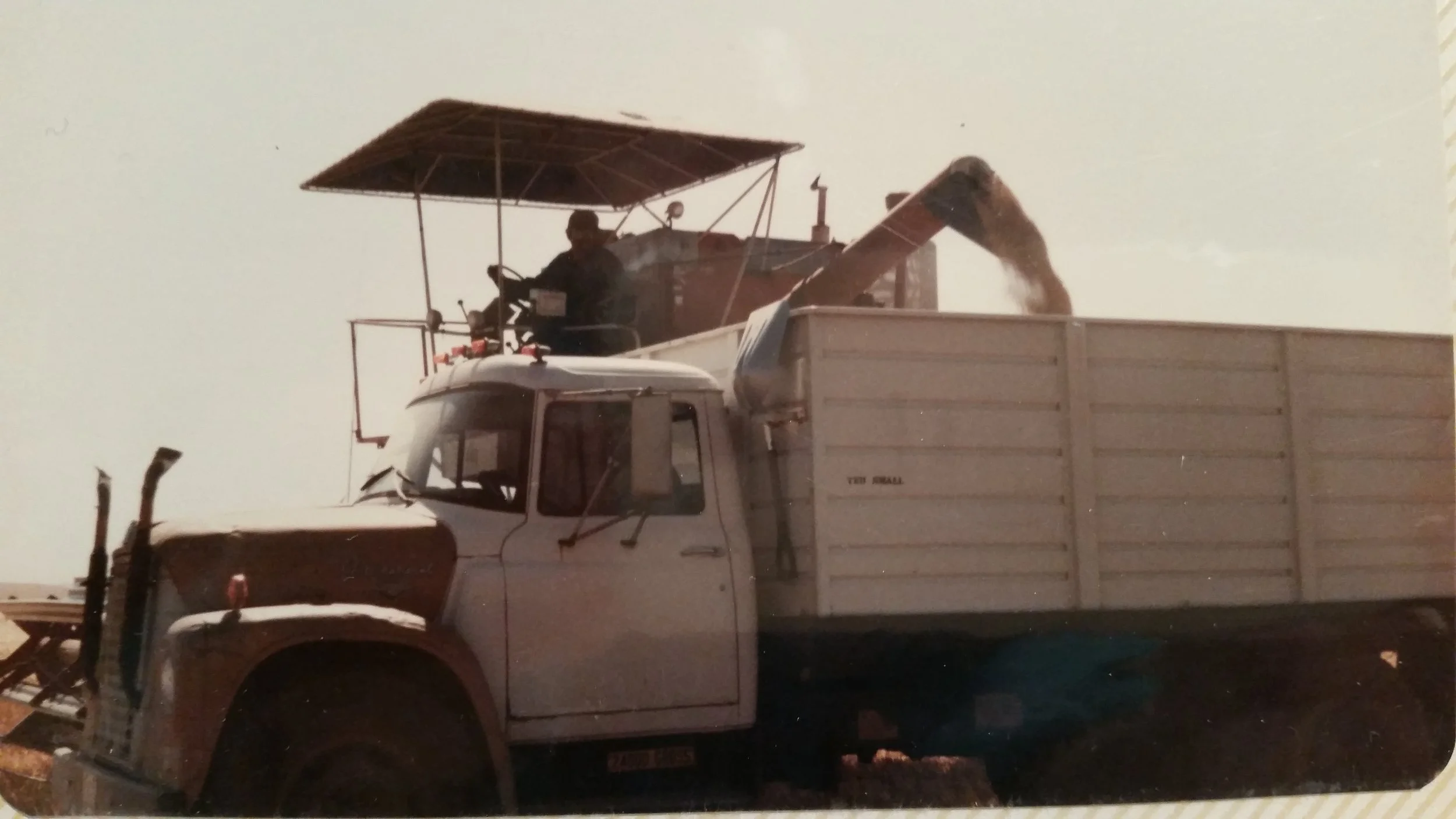 A combine with an augur pouring grain into a truck bed, seen against a foggy (maybe smokey) sky.