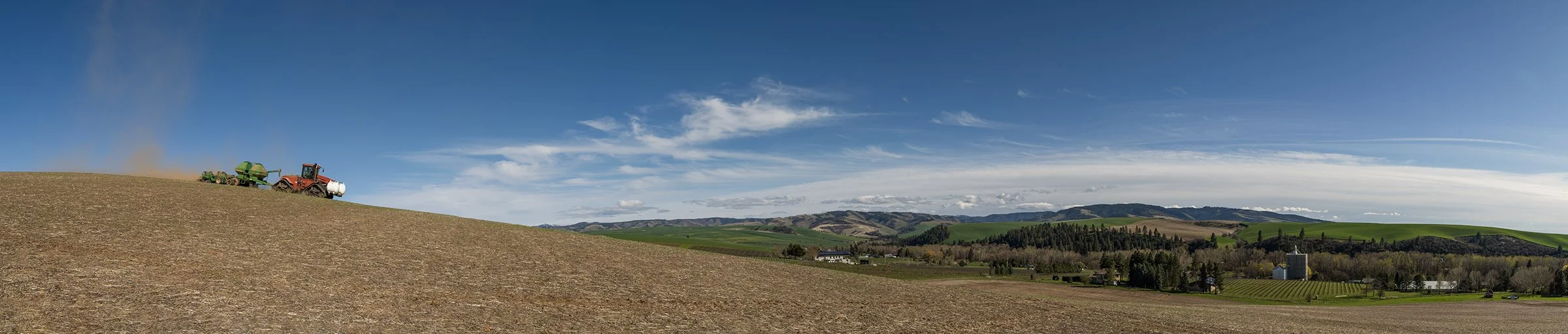 A panoramic view of rural farmland with a tractor and equipment on a hill, rolling green hills, trees, and a few buildings under a partly cloudy sky.