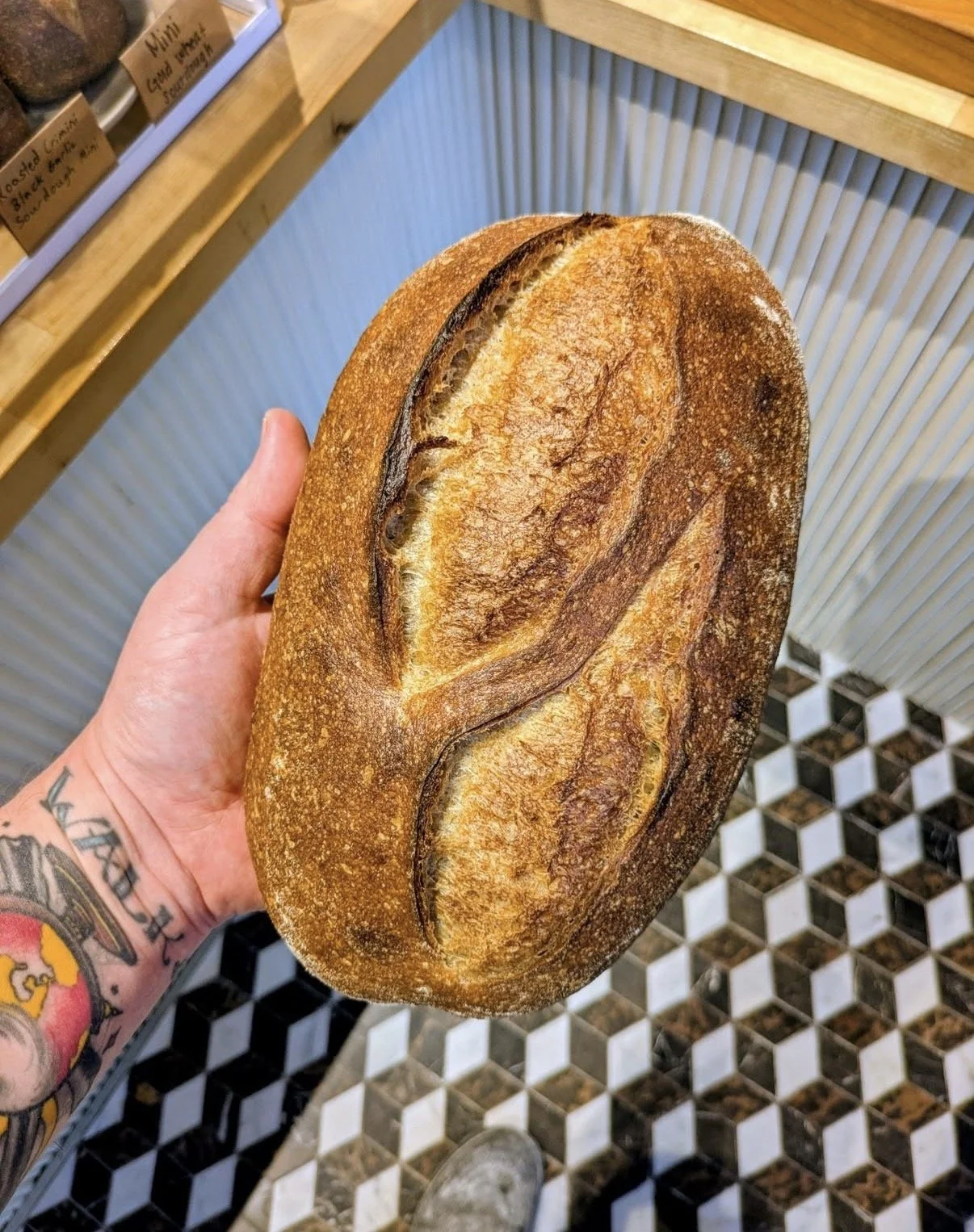 A person's hand holding a freshly baked loaf of bread with a crispy, golden-brown crust in a bakery setting.