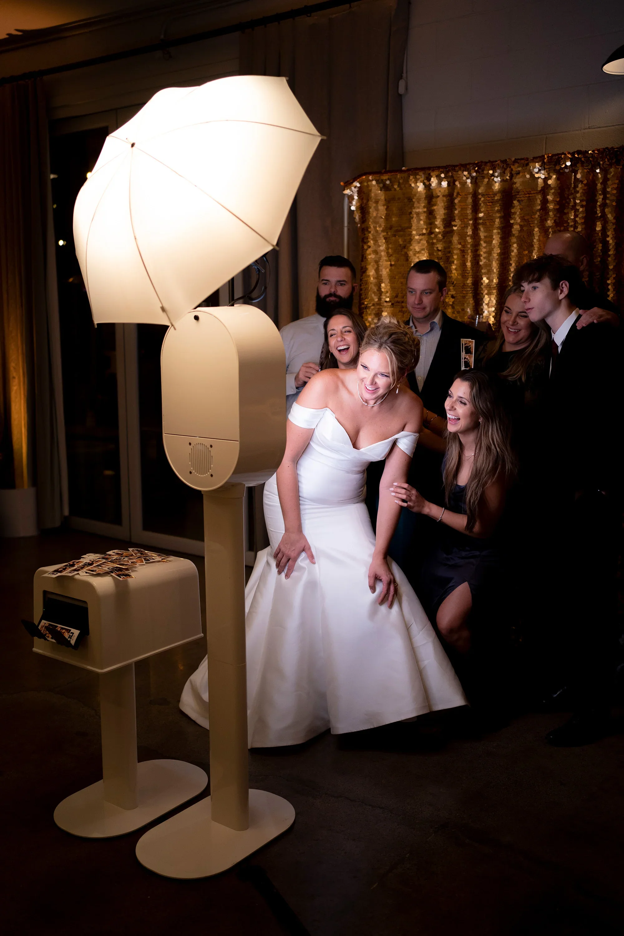 A group of six people, including a bride and groom, posing for pictures at a wedding reception with a floral backdrop and professional lighting.
