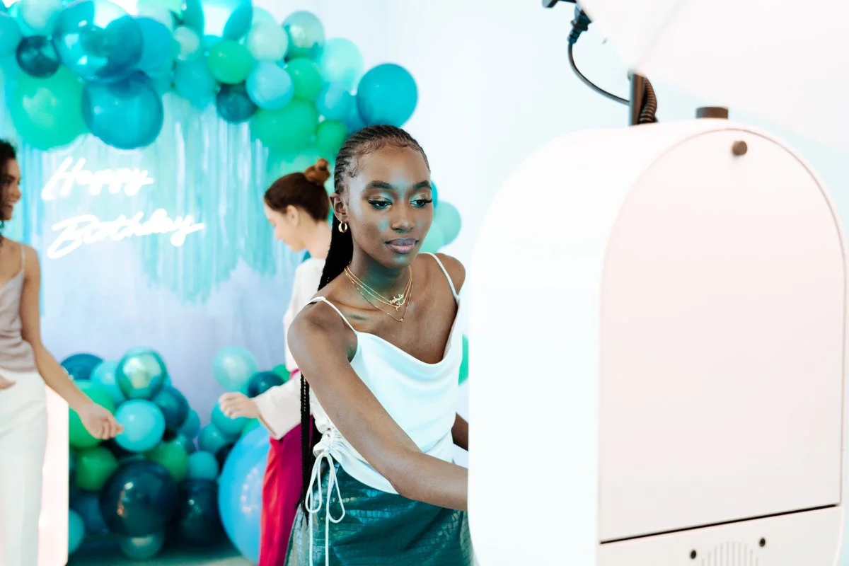 A woman at a birthday celebration taking a photo with a photobooth, with balloons and a "Happy Birthday" sign in the background.