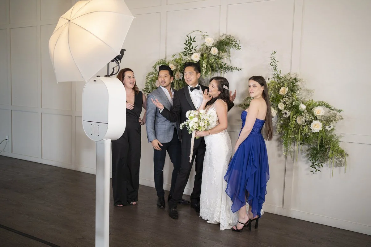 People taking a wedding photo in a studio with a large white umbrella light and a floral backdrop.