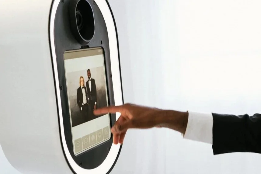 A person in a suit touching a touchscreen photo booth displaying a picture of a man and woman in formal attire.