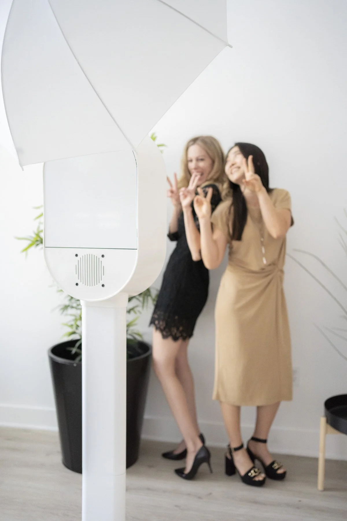 Two women standing in front of a photo booth, smiling and making peace signs, with a large umbrella light overhead.