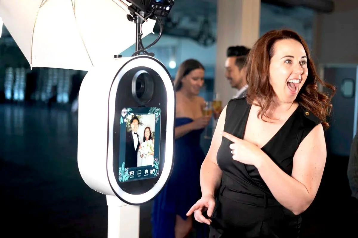 A woman with red hair in a black dress pointing and smiling at a photo booth camera that displays a picture of a recently married couple.