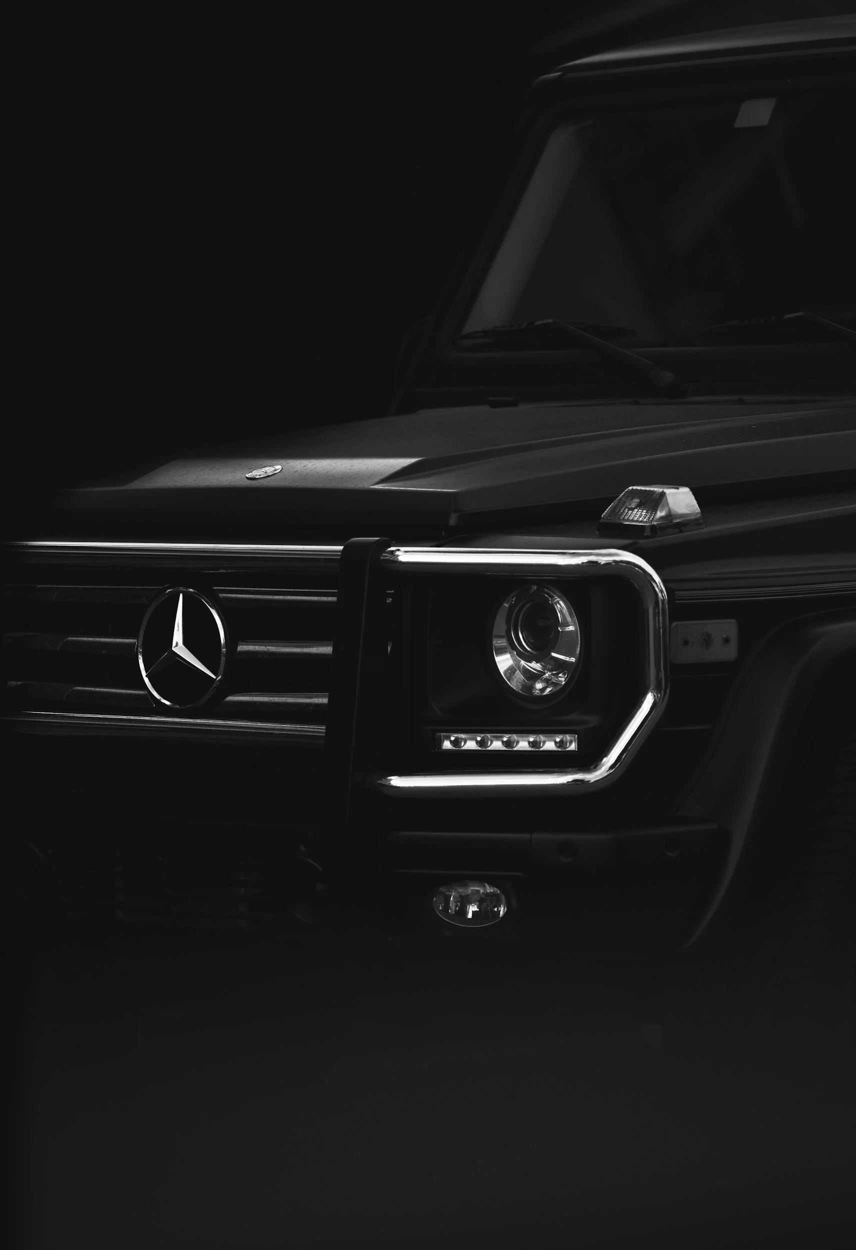 Close-up of the front of a black Mercedes-Benz SUV with distinctive grille, headlight, and emblem, partially illuminated.