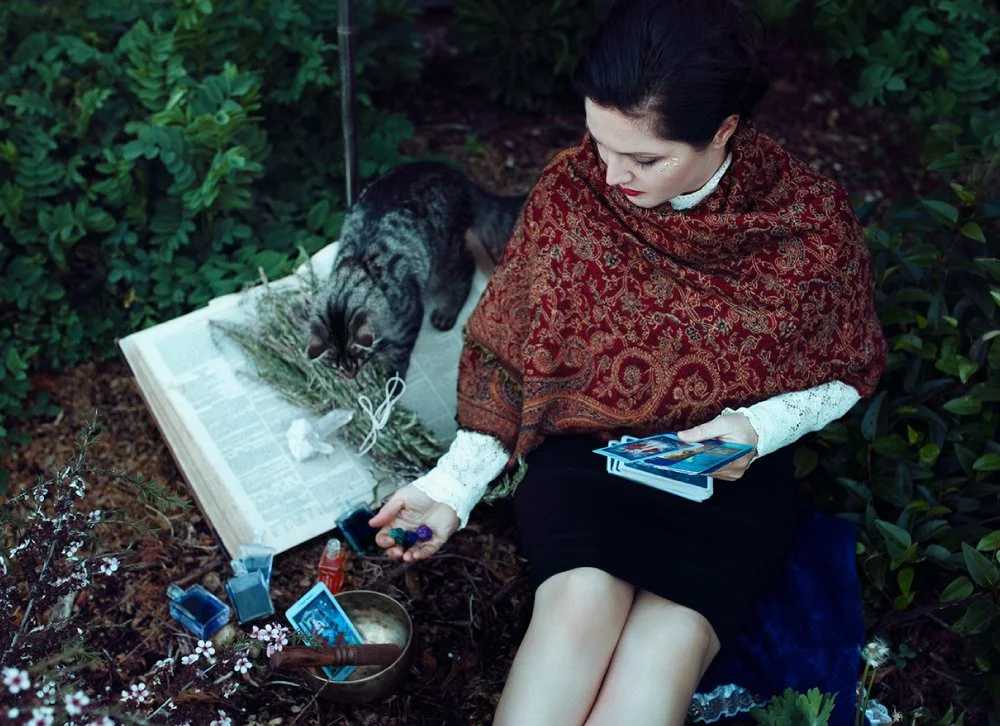 A woman with dark hair in a bun, wearing a red patterned shawl and white lace sleeves, sits outdoors surrounded by greenery. She holds tarot cards and a small jar. A black and grey cat is lying on an open book nearby, which has herbs and string on it. There are small bottles, cards, and a bowl in front of her.
