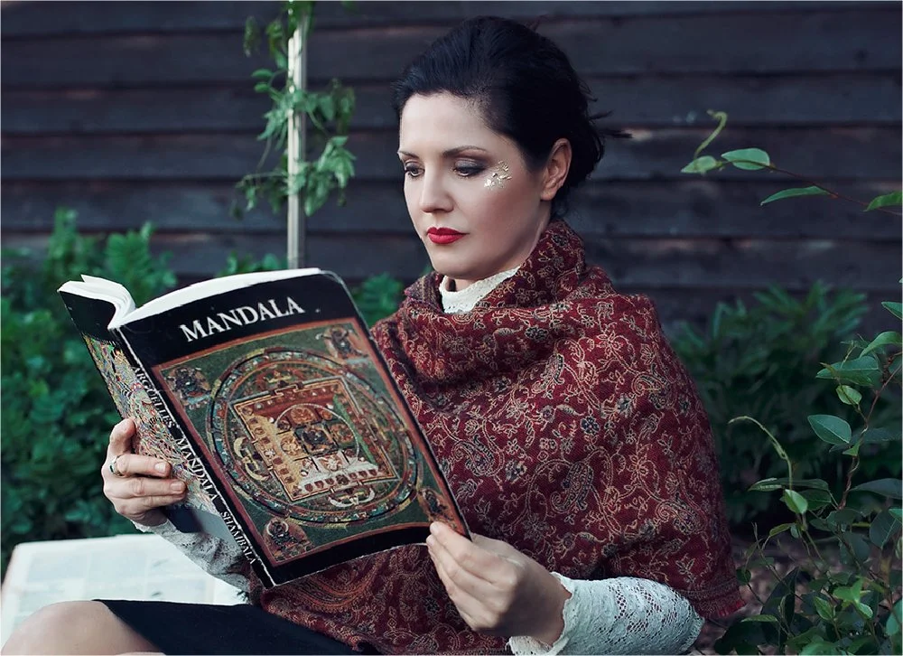 Woman with dark hair, wearing a red and gold patterned shawl and white lace gloves, reading a book titled 'Mandala' outdoors near a wooden fence and green plants.