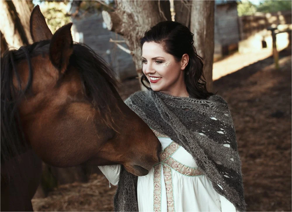 A woman with dark hair smiling and looking at a brown horse outdoors in a wooded area, wearing a gray shawl over a white dress.
