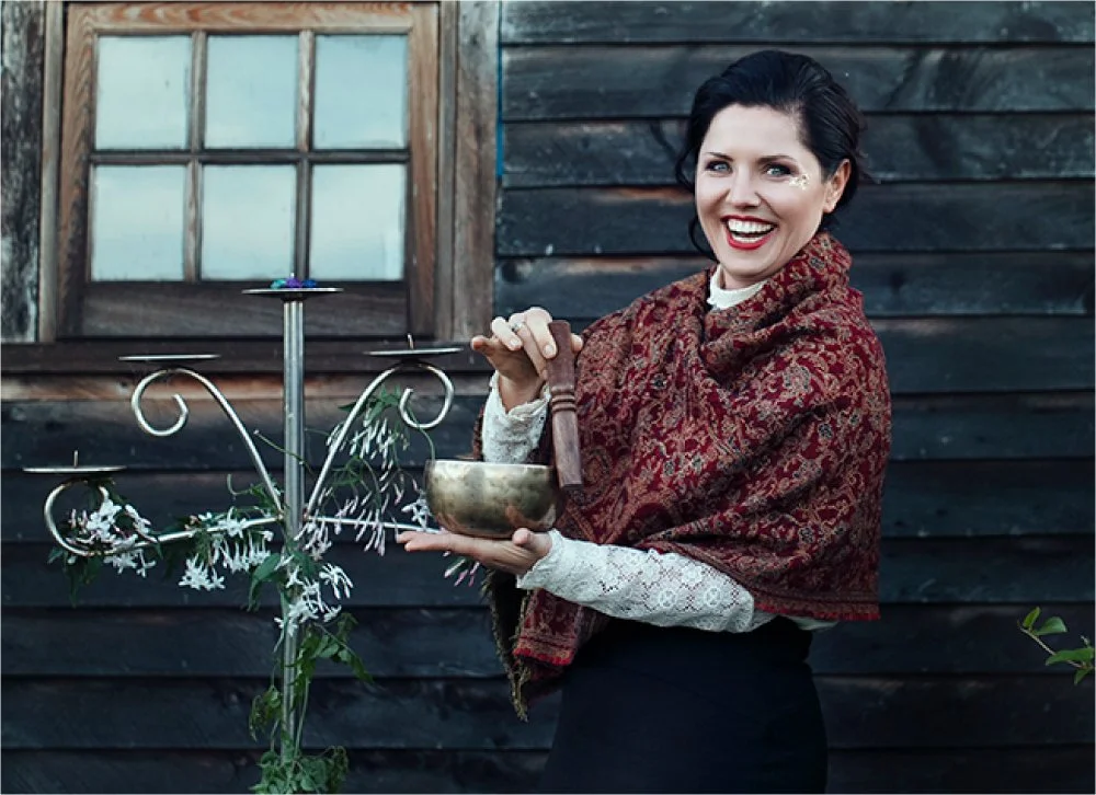 A woman with dark hair, wearing a patterned red shawl and lace sleeves, smiling and holding a singing bowl and mallet outdoors in front of a dark wooden building with a window.