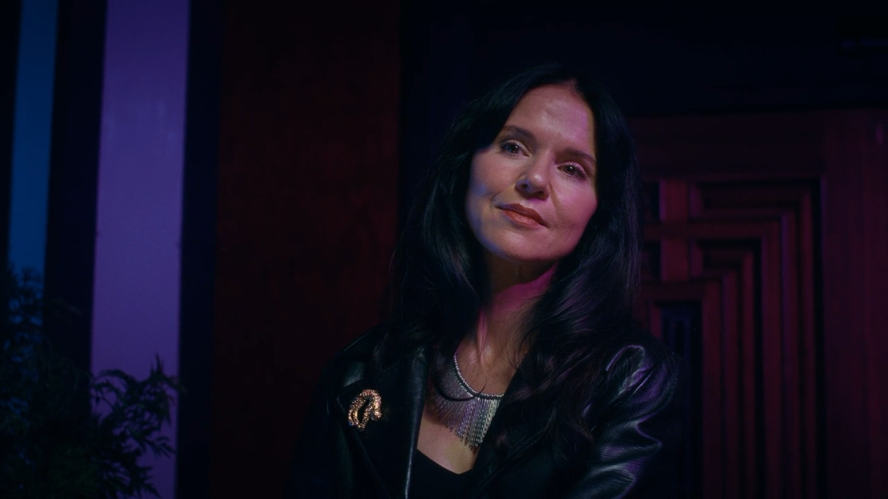 A woman with long black hair wearing a black leather jacket and silver jewelry, standing indoors with colorful lighting.