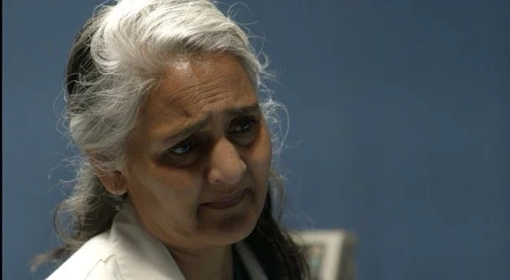 Close-up of an elderly woman with silver hair tied back, showing a concerned or confused expression.