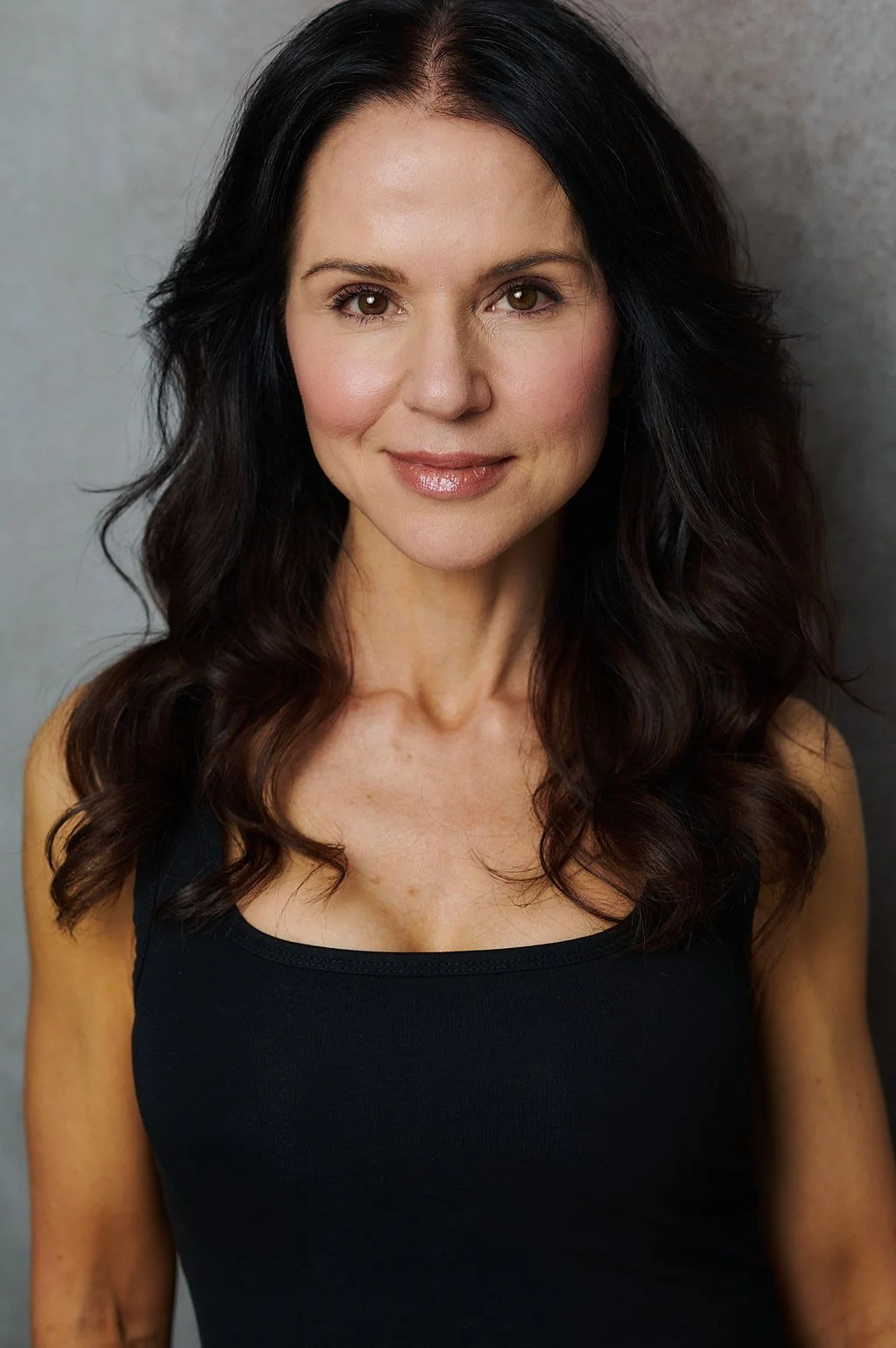 A woman with long, dark, wavy hair wearing a black tank top, smiling softly, against a plain background.