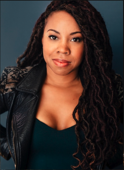 A woman with long, curly hair wearing a black leather jacket and a dark top, posing against a neutral background.