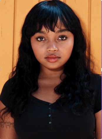 Young woman with dark hair and bangs, wearing a black top, standing against a yellow wall.