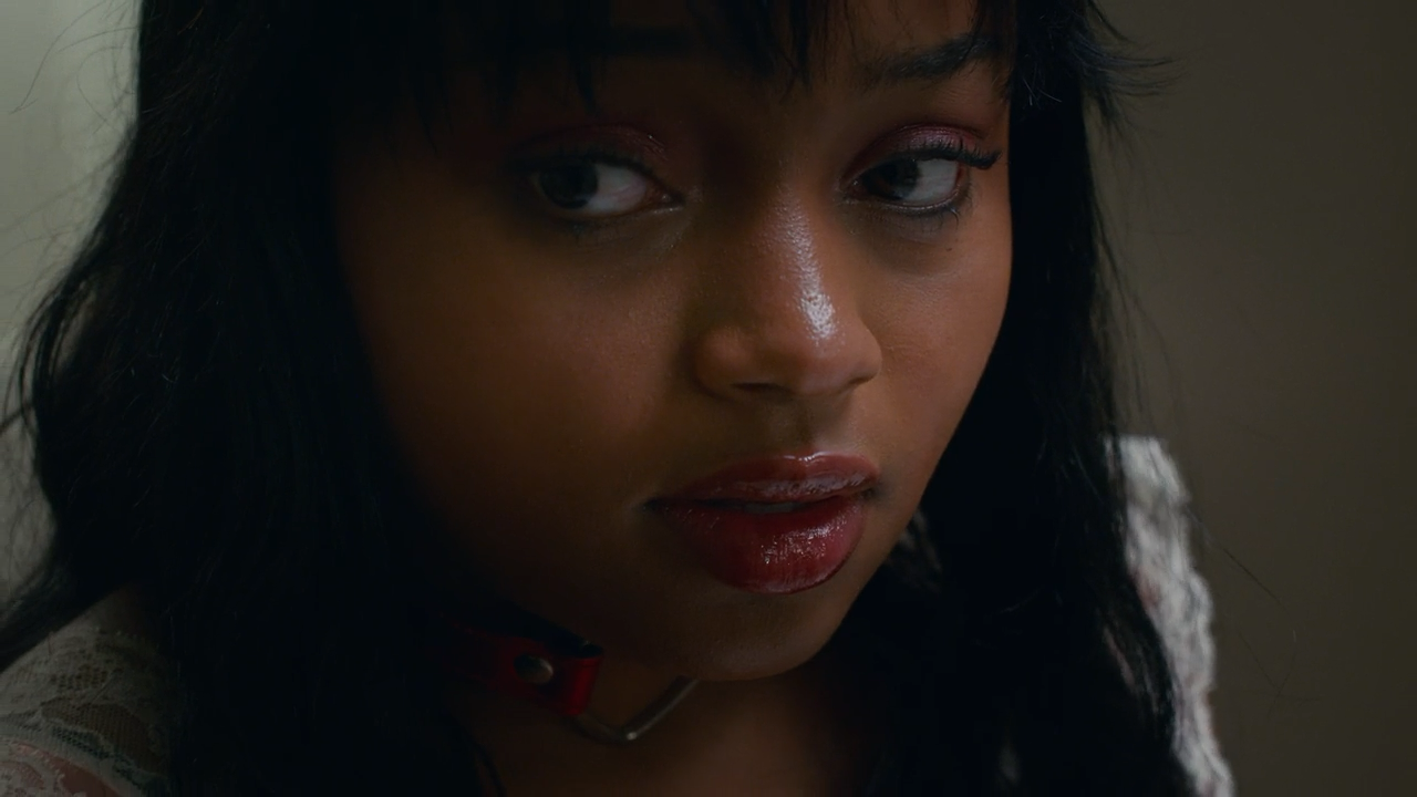 A young woman with dark hair and makeup looking down and to the side, wearing a red collar and lace clothing.