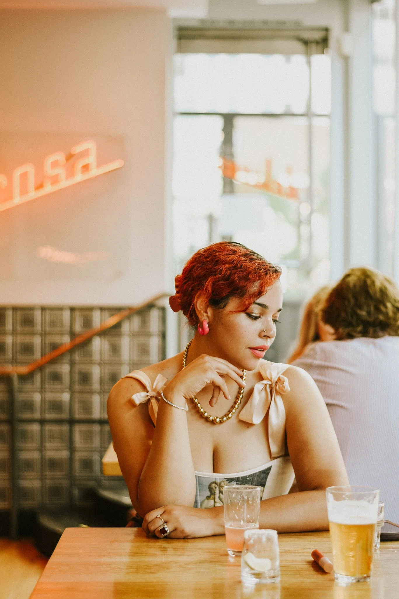 A woman with red hair sits at a wooden table in a cafe, wearing a beige dress with bow sleeves, pink earrings, a pearl necklace, and various jewelry. There are drinks on the table in front of her and she appears to be looking down thoughtfully.