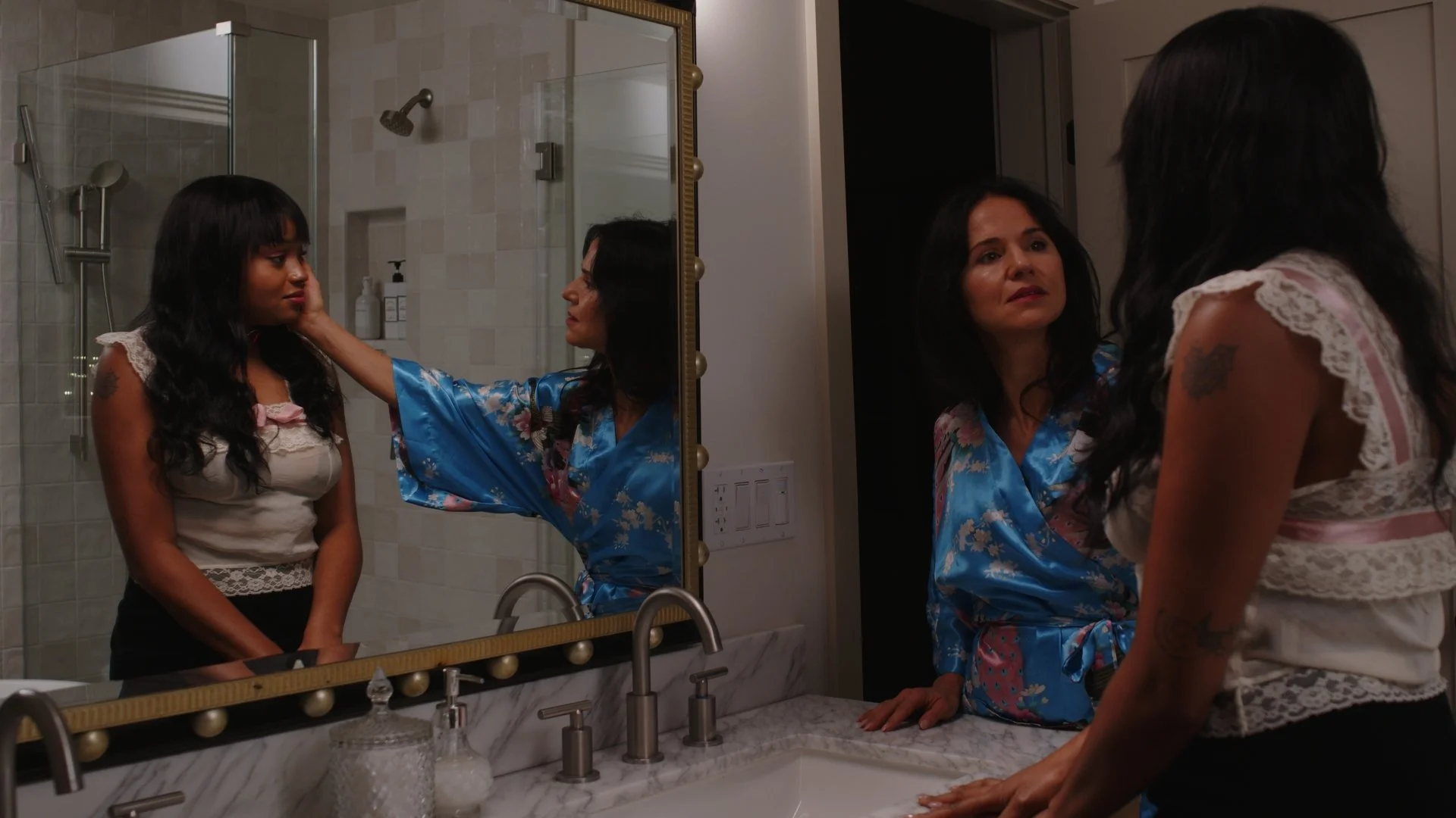 Two women looking at each other in front of a mirror in a bathroom. One woman is looking at herself, while the other woman touches her face, with the shower and toiletries visible in the background. The woman touching the face has dark hair and is wearing a blue satin robe with floral patterns. The woman she is touching has dark hair and is wearing a sleeveless top with lace details.