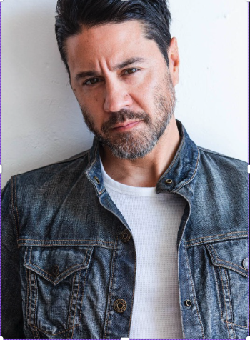 A man with dark hair and a beard leaning against a wall, wearing a denim jacket over a white shirt, looking seriously at the camera.