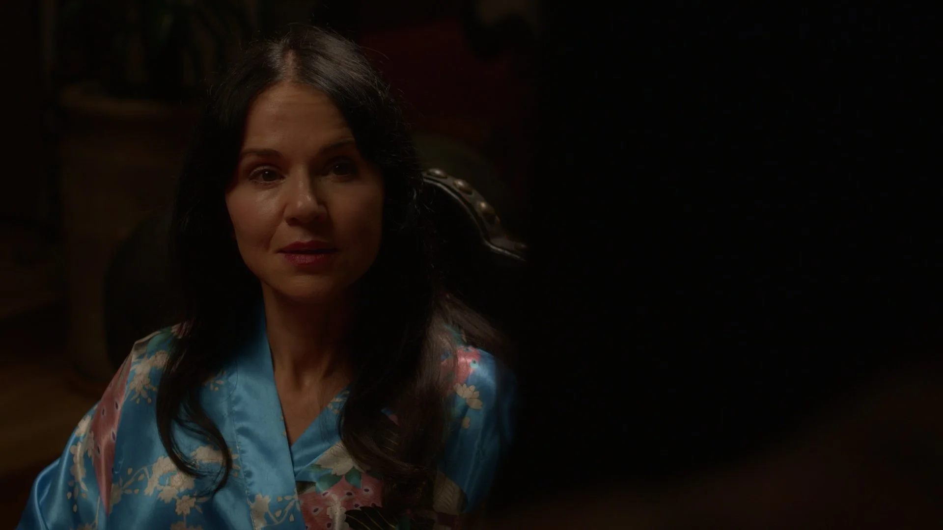 A woman with dark hair sitting in a dimly lit room, wearing a blue floral dress, looking ahead with a neutral expression.