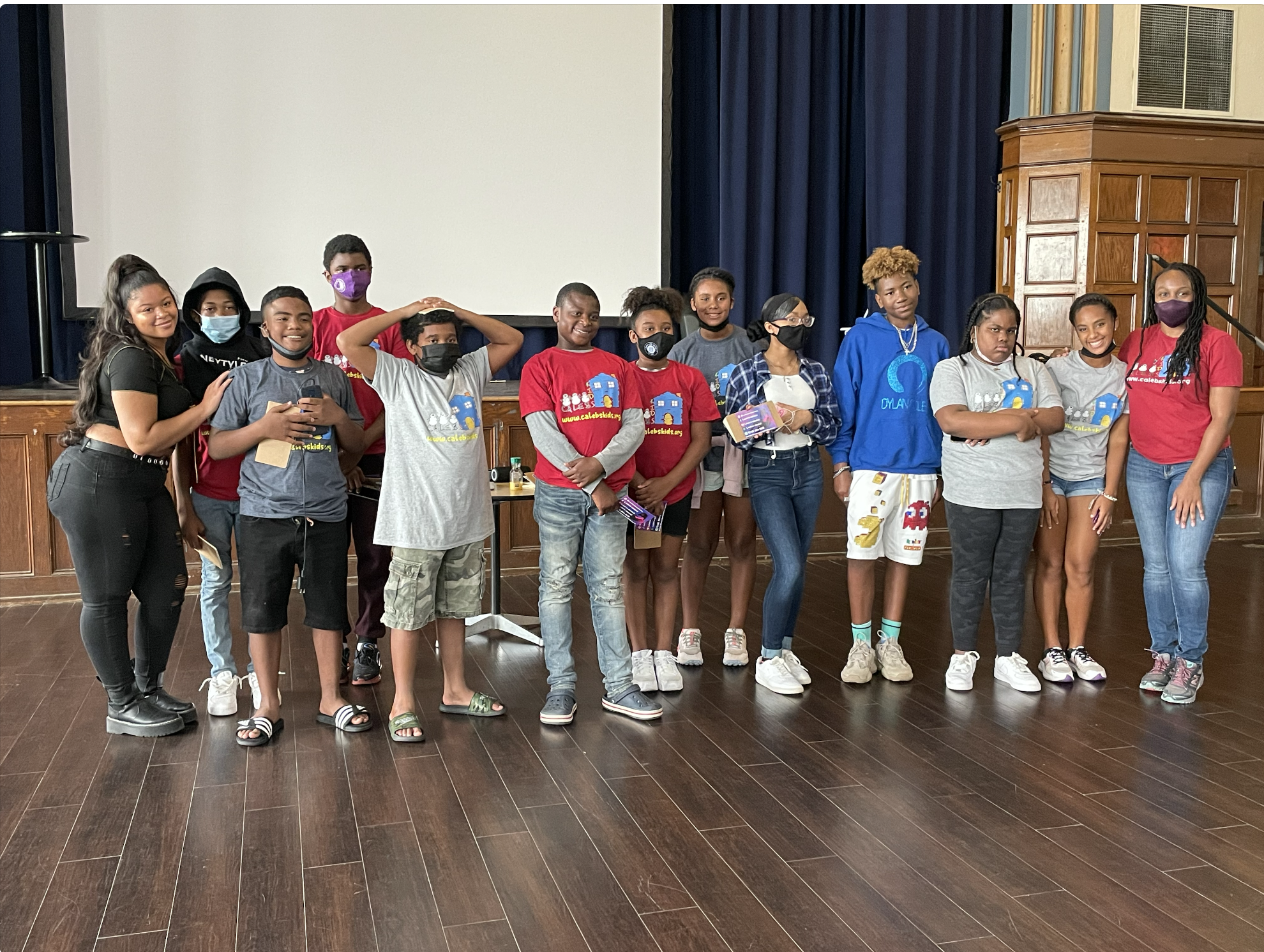 A group of young diverse children and two women standing on a stage in a hall, some wearing masks and T-shirts, posing for a photo during an event.