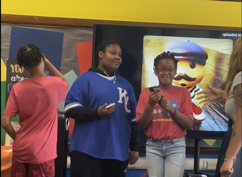 A group of children in a classroom with colorful decorations. Two girls are standing near a large screen; one girl in a red shirt is holding a microphone, smiling, and the other girl in a blue shirt with white lettering is holding a phone. A boy in a pink shirt is standing with his back to the camera.