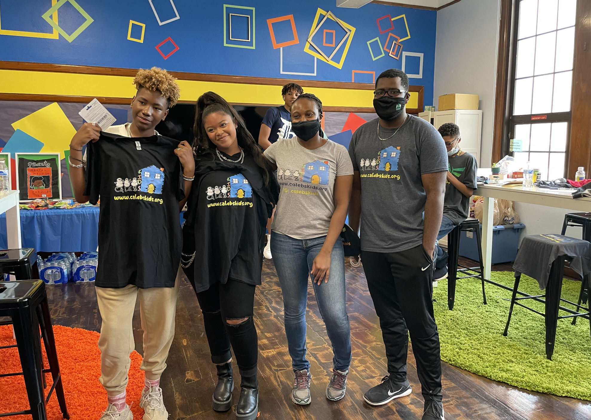 Group of five diverse people in a room with colorful geometric decorations, some standing and some sitting, wearing Calvin Klein t-shirts and face masks.