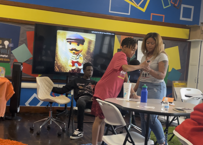 Three children in a colorful room, one sitting on a chair, another standing and showing a phone to a girl, with a large screen displaying a cartoon character in the background.