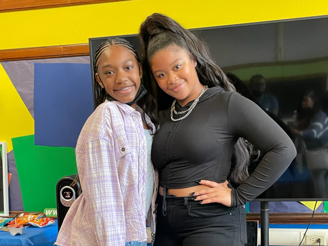 Two young women posing indoors, one with long straight hair and the other with braided hair, smiling at the camera, with a television behind them.