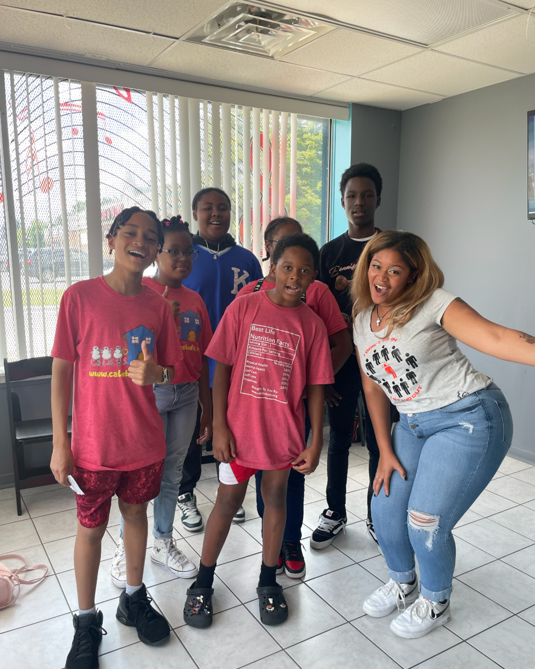 Group of seven children and one adult woman indoors, smiling, posing for the photo, with a window and blinds in the background.