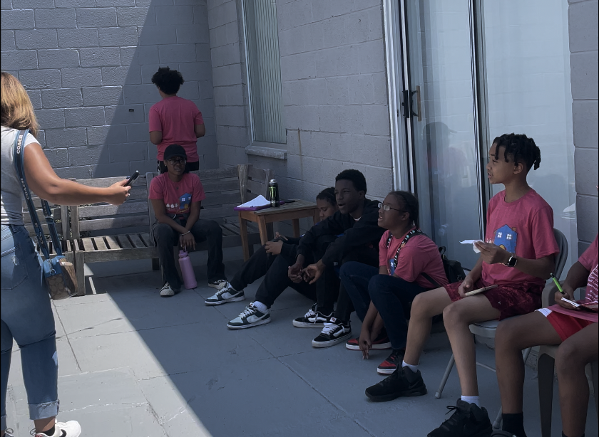 Group of children sitting and standing outside, with some taking notes and one person recording, on a patio with a brick wall and sliding glass door.