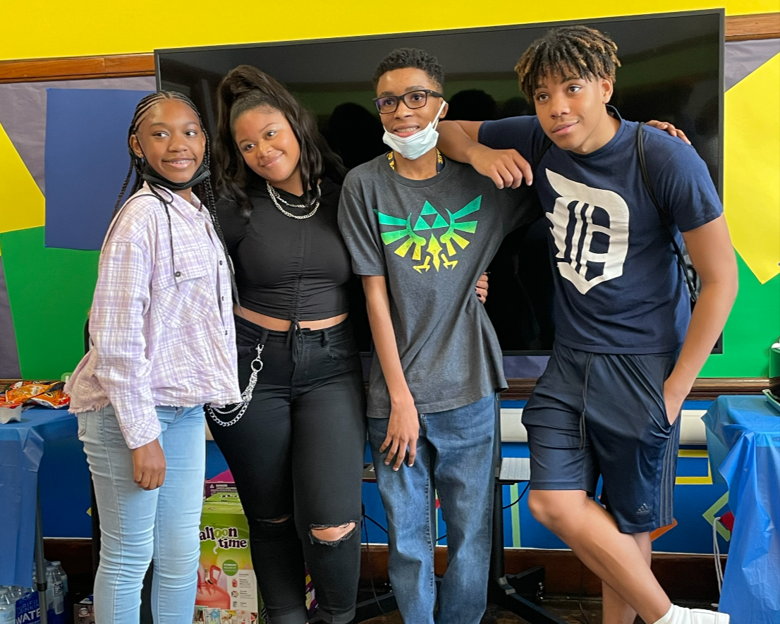 Four teenagers standing together indoors in front of a large black screen, smiling and posing for a photo.