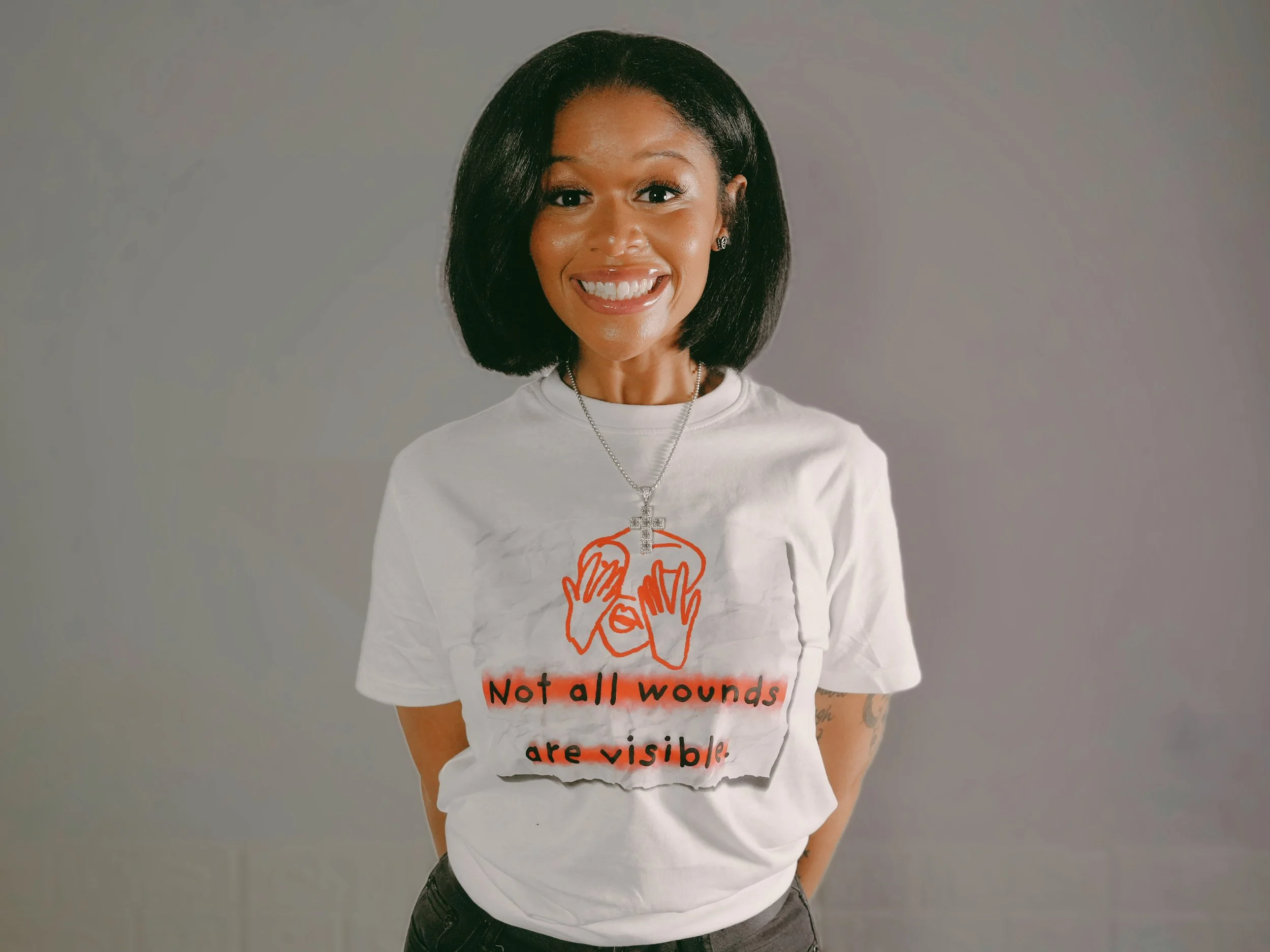 Woman smiling, wearing a white t-shirt with a red illustration of hands covering eyes and the text 'Not all wounds are visible', standing against a plain background.