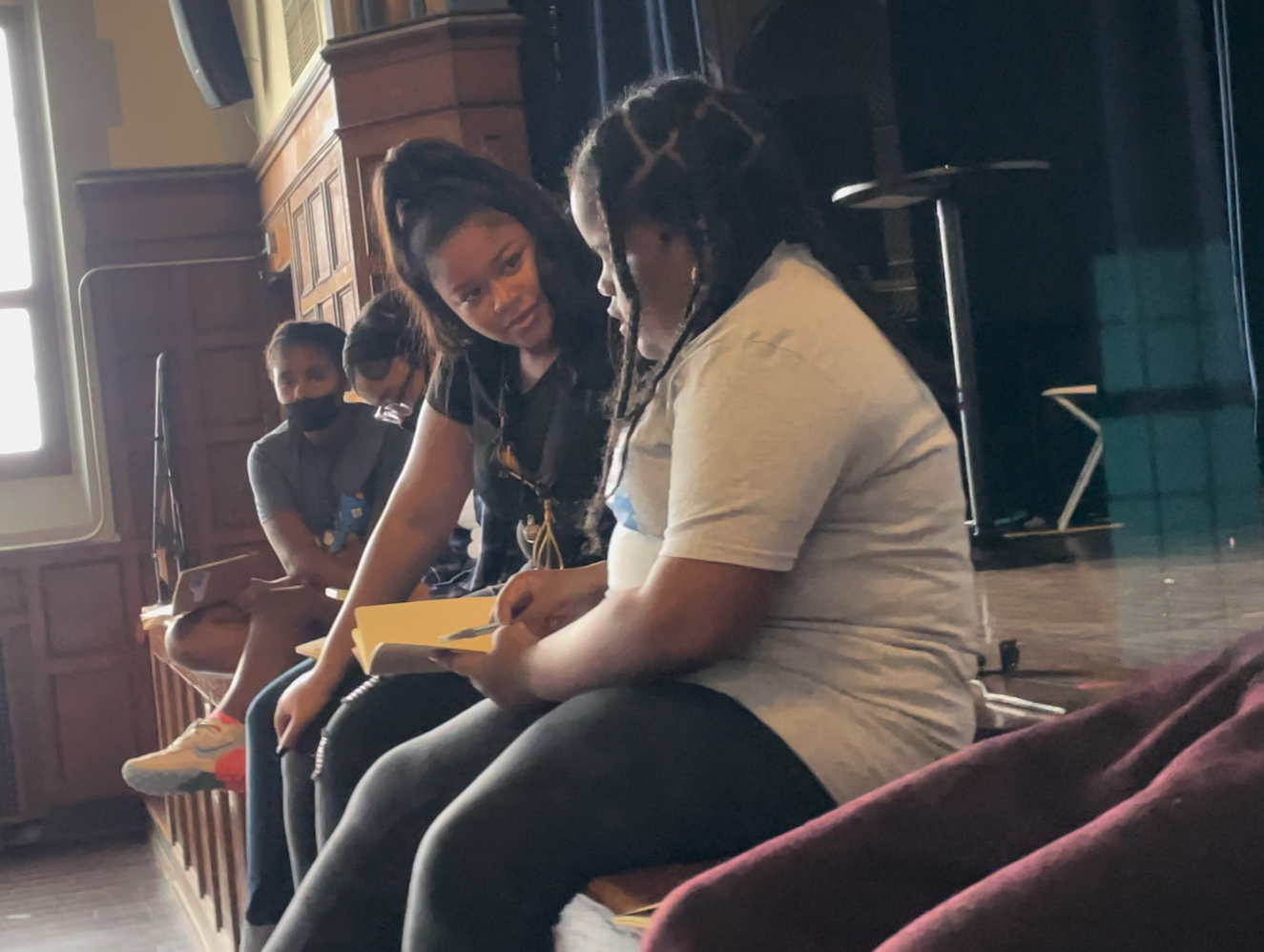 A group of children sitting on a bench, with the two girls in the foreground having a conversation, one of whom is holding a yellow notebook. The children are in a room with wooden paneling and a large window. Some of the children are wearing face masks.