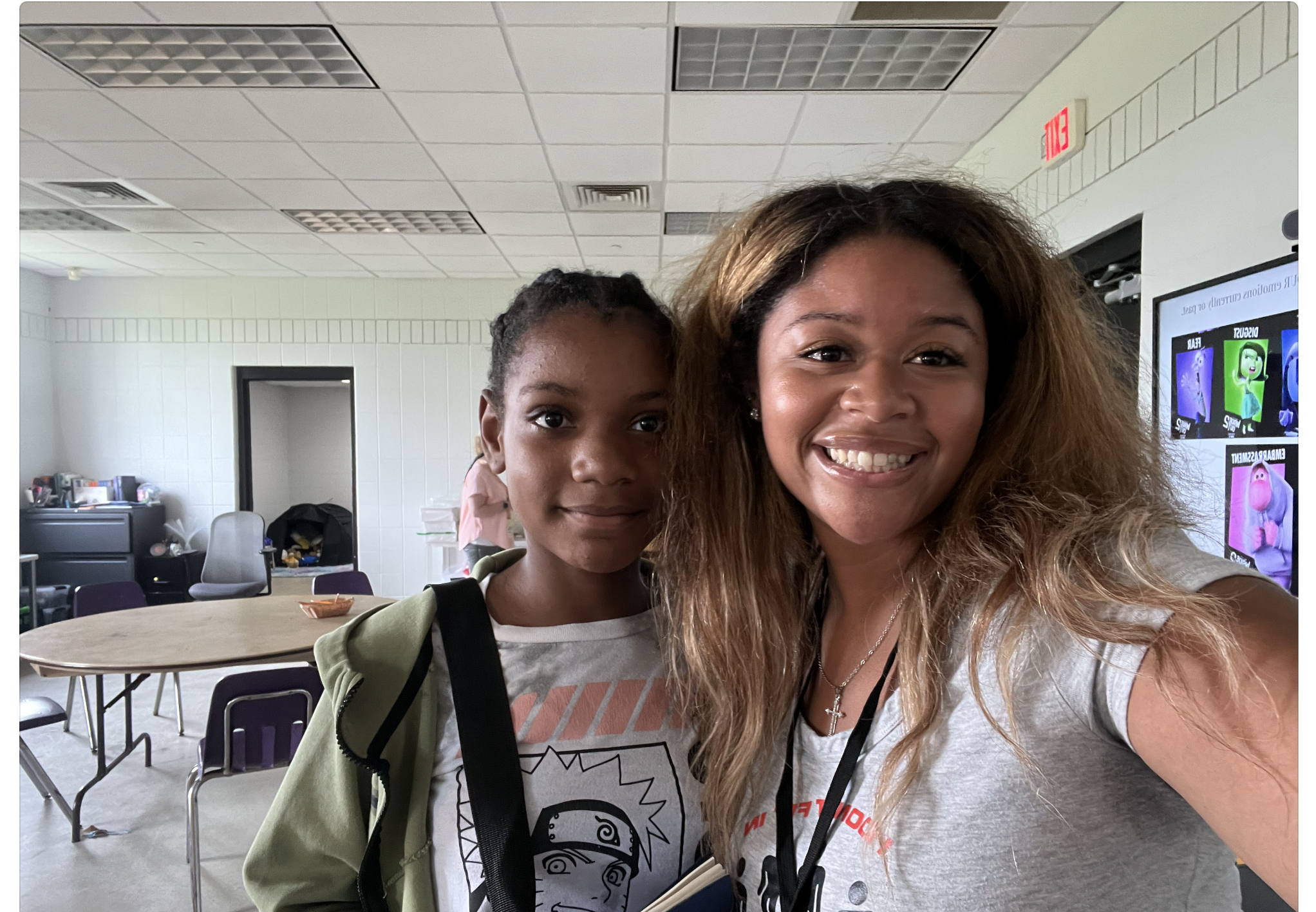 Two smiling women taking a selfie in a room with tables, chairs, and a screen displaying animated characters in the background.