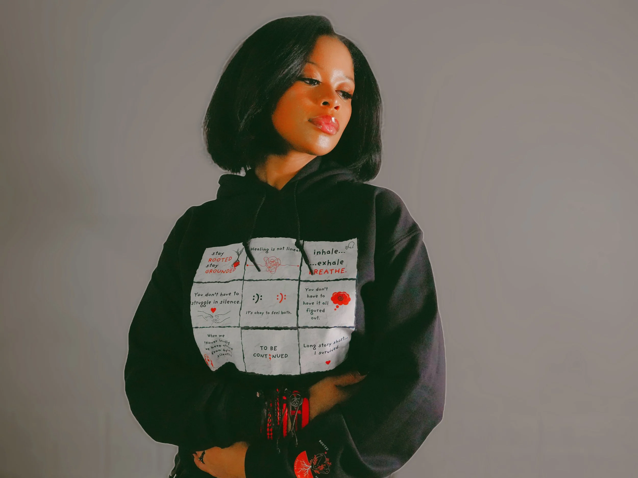 A young woman with shoulder-length black hair, wearing a black hoodie with a printed design, stands against a plain gray background. She looks slightly to the side with a confident expression.