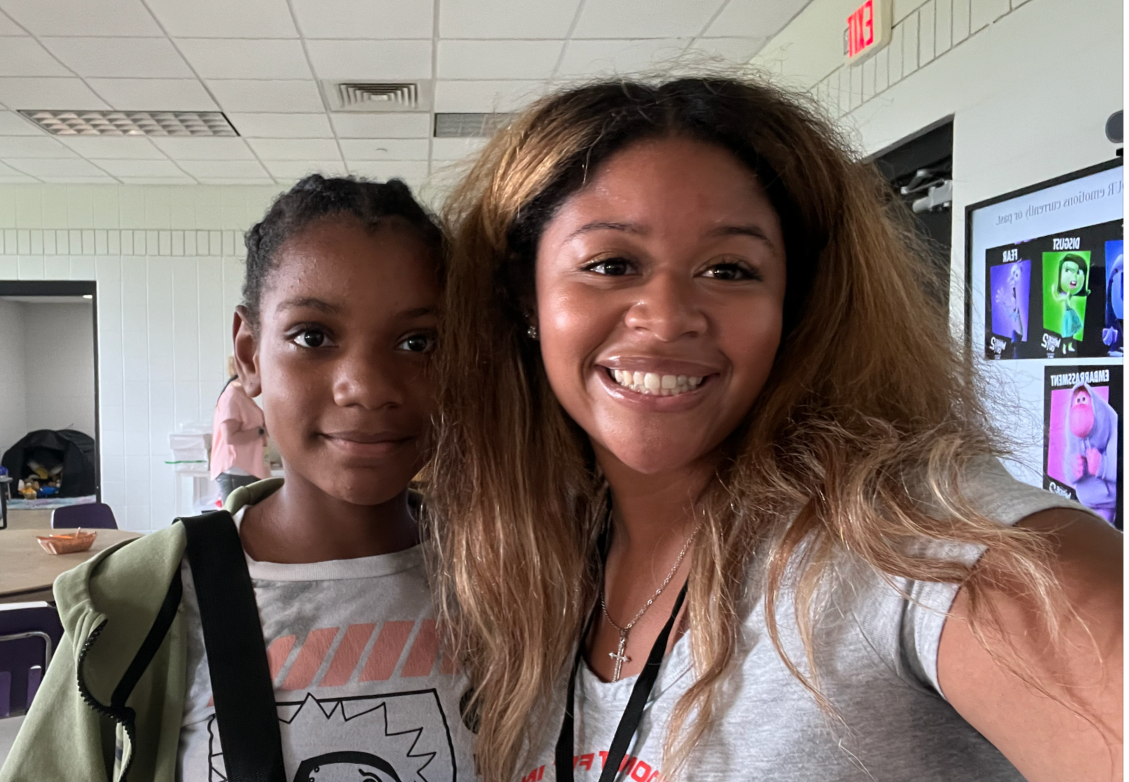 A woman and a young girl taking a selfie together in a classroom or conference room. The woman has blonde, wavy hair and is smiling widely. The girl has dark hair styled in braids and is also smiling subtly. In the background, there is a digital screen displaying colorful cartoon characters.