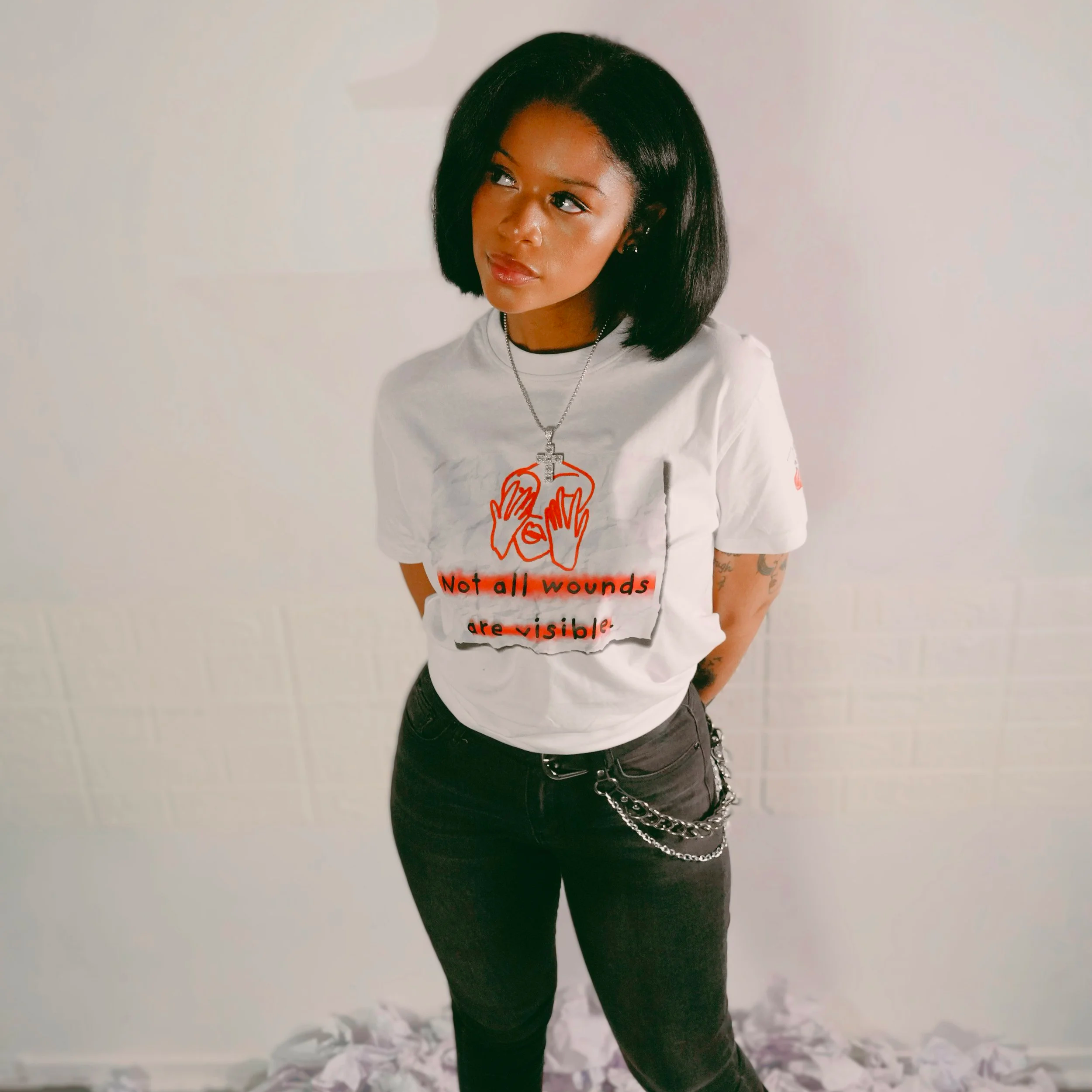 A woman with short black hair wearing a white T-shirt with red text and hands illustration, paired with black jeans and a chain accessory, standing against a plain light background.