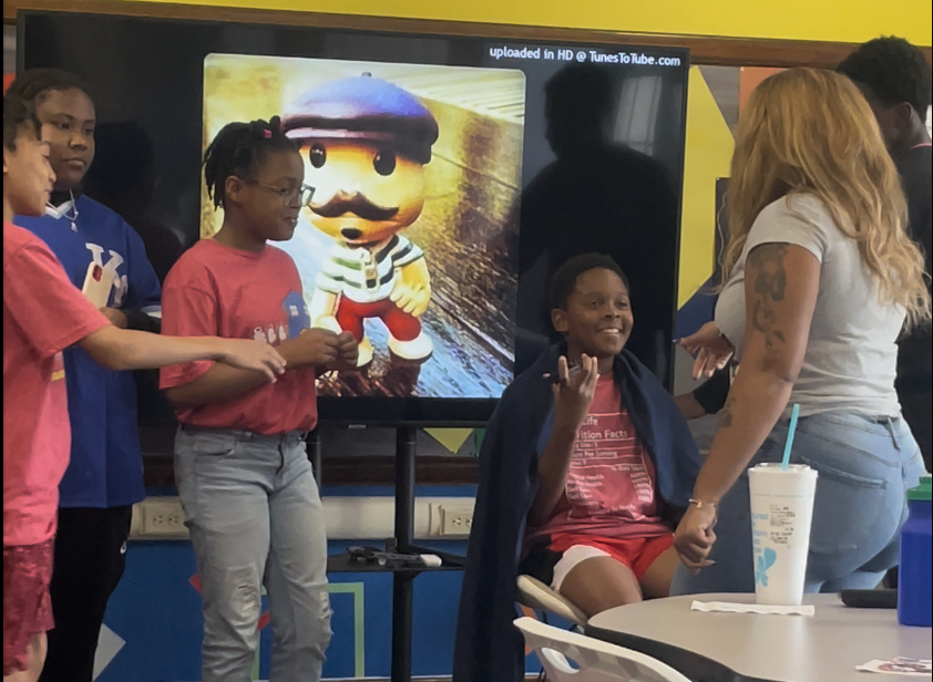 A group of children and a woman in a classroom, with a large screen showing a cartoon character and children gathered around.